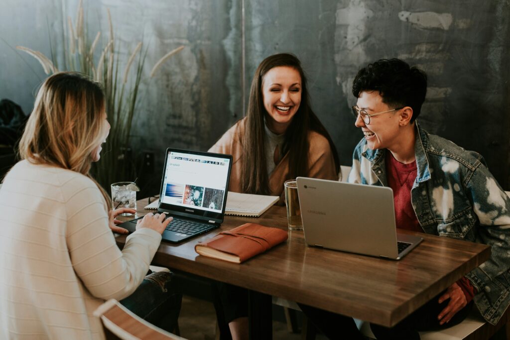 Three TEFL course students laughing and studying together with laptops at a café table abroad
