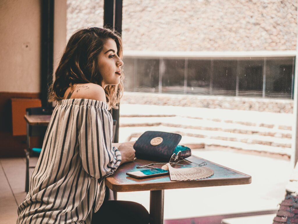 Woman sitting alone at a café table looking out the window with a distant expression, experiencing reverse culture shock after returning home from abroad