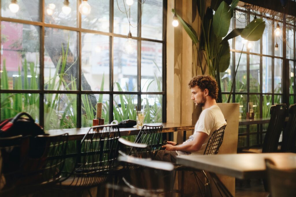 Man sitting alone at a café table by a large window, looking thoughtful and distant