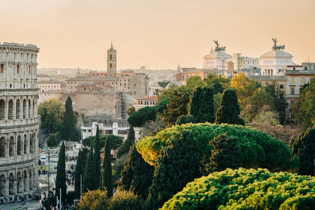 Aerial view of Rome at golden hour with the Colosseum in the foreground, cypress trees, and the Altare della Patria visible in the distance