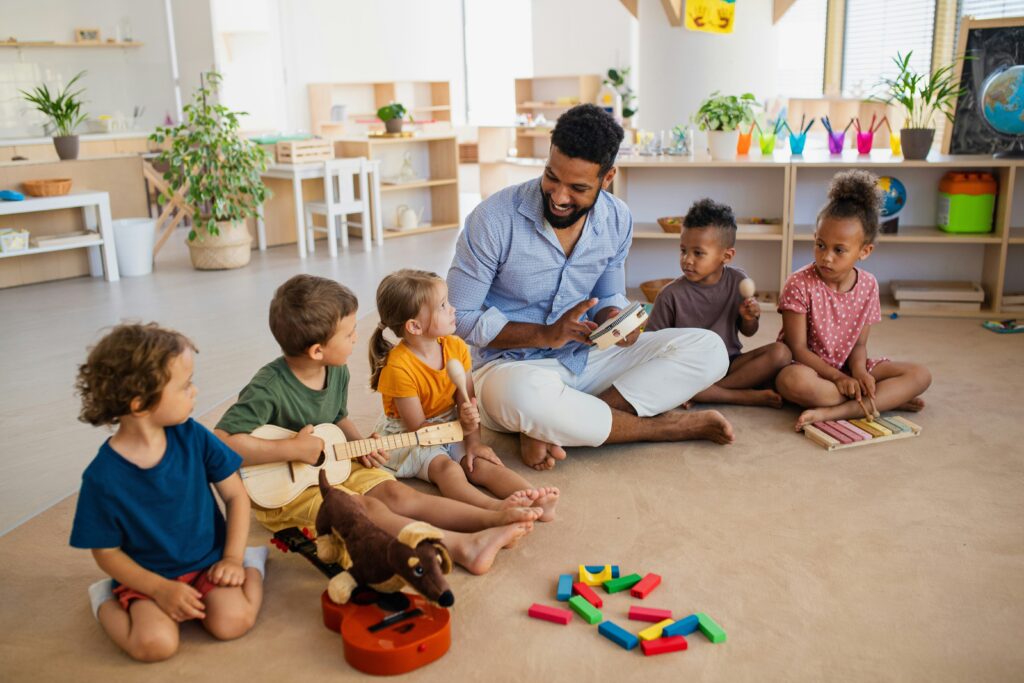 Male teacher sitting on the floor with young children playing instruments in a bright classroom — the kind of first teaching position accessible to those who want to teach abroad with no experience