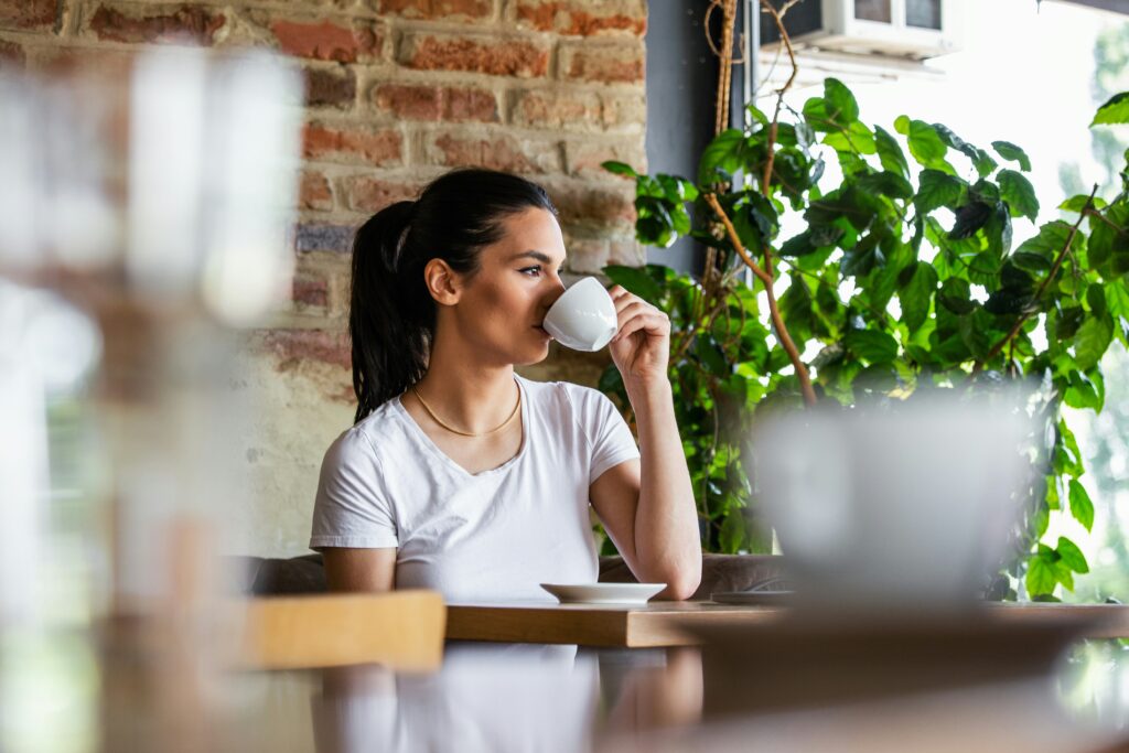 Woman sitting alone at a café table drinking coffee and looking out the window, lost in thought after returning home from living abroad