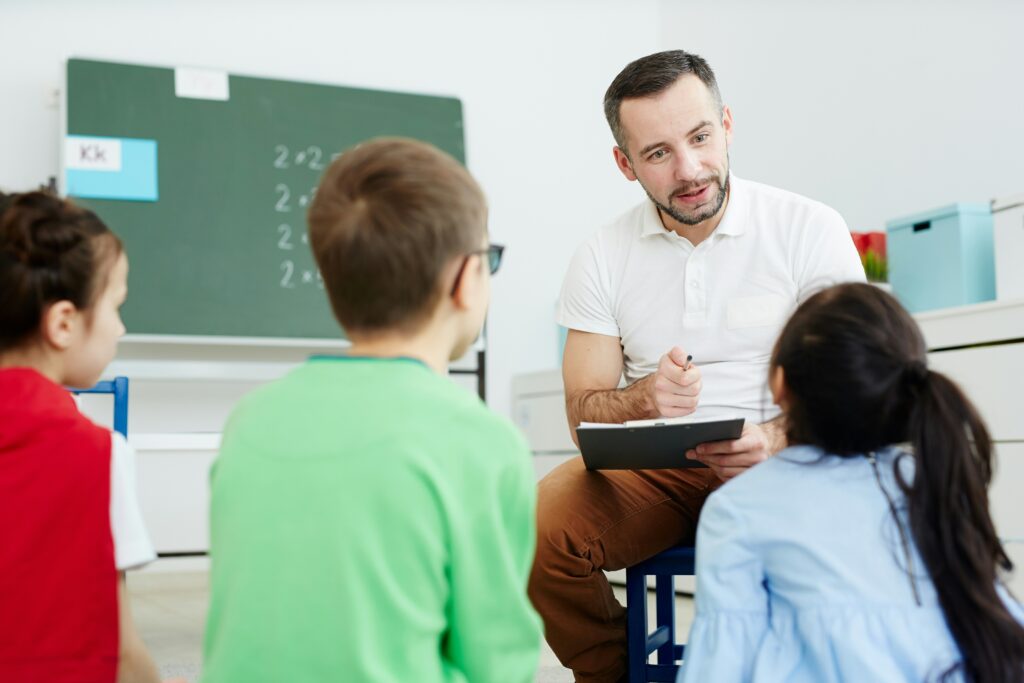 Male English teacher sitting with small group of young students in a classroom abroad answering teaching abroad FAQs