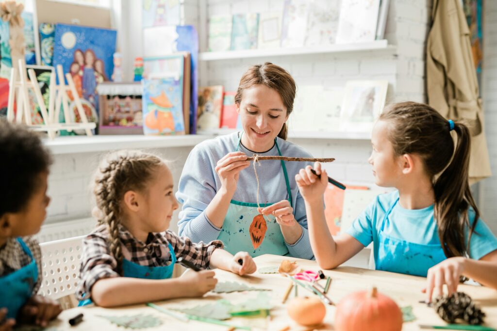 Female teacher in an apron engaging with young students at a craft table in a colorful classroom — the type of hands-on teaching environment awaiting those who teach abroad with no experience