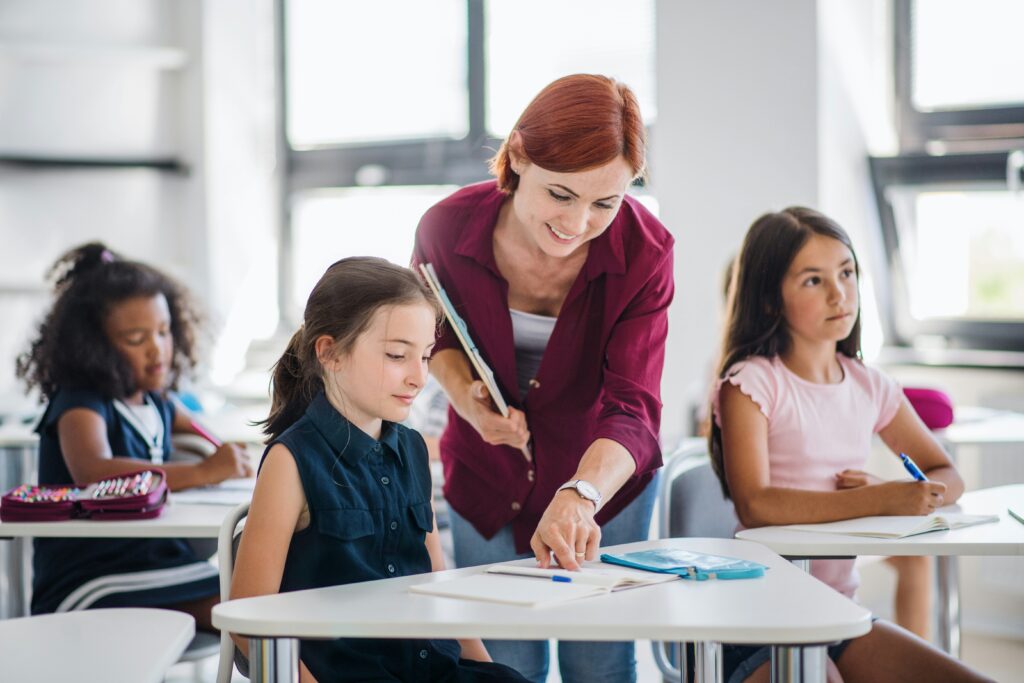 Female English teacher helping a young student at her desk in a bright classroom abroad