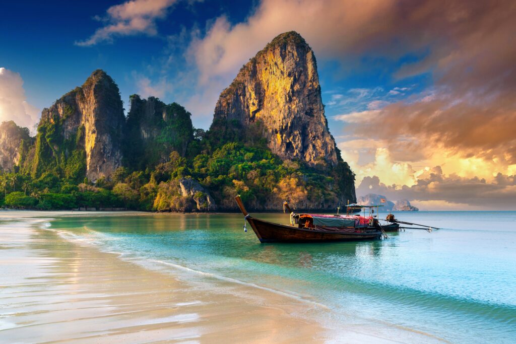 Longtail boat on the white sand shore of Railay Beach in Krabi, Thailand — one of the best weekend destinations for teachers who teach English in Thailand — with dramatic limestone karst cliffs at sunset
