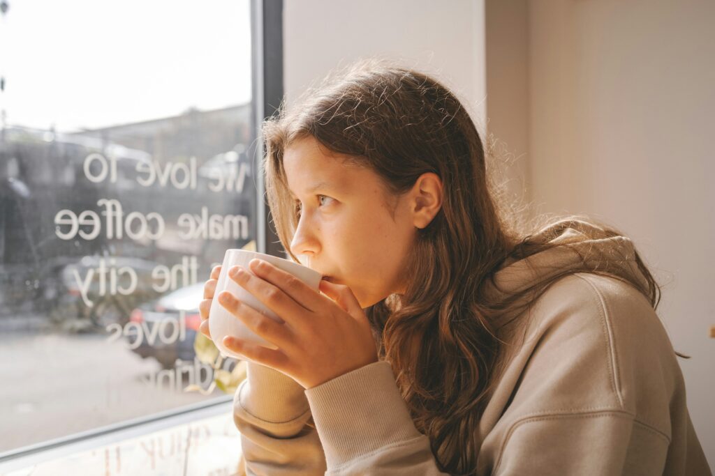 Young woman sitting alone in a café holding a coffee cup and looking out the window with a distant expression — capturing the quiet loneliness that what nobody tells you before you teach abroad warns you about