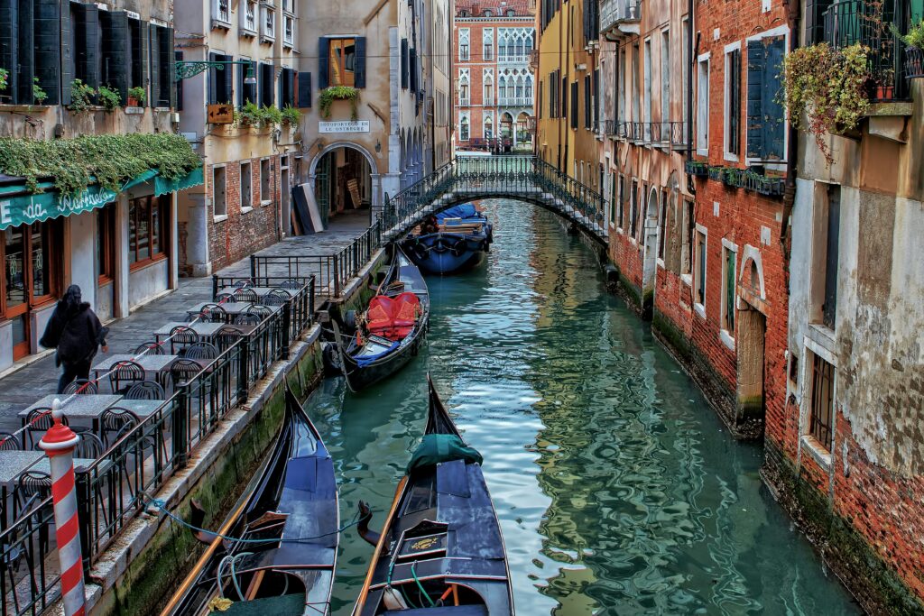 Narrow canal in Venice, Italy with gondolas moored alongside brick buildings and a small stone bridge in the background. Scenic view for teaching abroad in Italy.