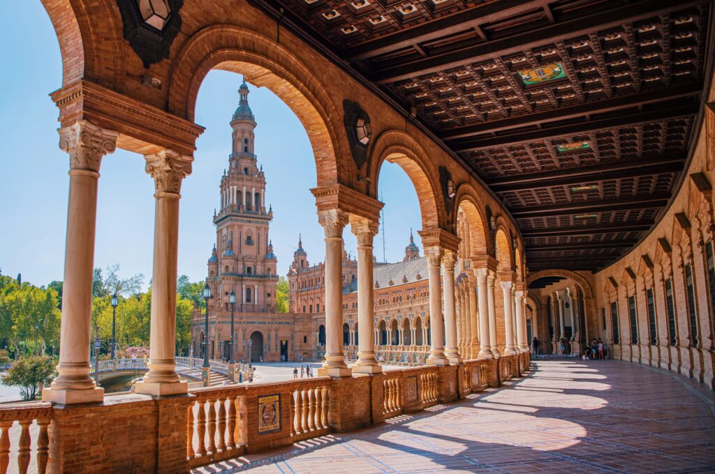 The ornate arched colonnade of Plaza de España in Seville, Spain, with its tower and decorative tilework visible in the background on a sunny day